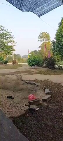 A dirt path leads away from the camera. On the left, there are piles of sand and what appear to be discarded shoes. In the middle of the path, there are some bricks, a pink plastic bag, and more debris. In the background, there are trees, a flagpole with flags, and what appears to be a group of people on a motorcycle traveling down the path. The sky is bright and clear.