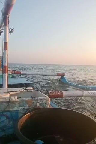 A video shows a view from a boat at sea. In the foreground, a large black container is visible. Behind it, there is a white container with wires and blue and white trim. A person sits on the left side of the frame, looking out at the water. Further out at sea, another boat with a blue and white hull and a mast is visible. The water is choppy, and the sky is clear with a soft, warm light, suggesting sunrise or sunset. Two poles with red and white stripes extend from the boat in the foreground towards the water.