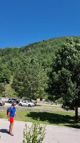 This video captures a scenic view of a rural landscape under a bright blue sky, suggesting a sunny day. The foreground shows a paved area with a red car parked on the right, and a white car on the left. Behind them, a grassy area is bordered by trees and a wooden fence. In the background, a lush green mountain rises steeply, covered in dense forestation. The scene appears to be a parking area or roadside near a natural park or mountainous region. There are several other vehicles visible, including a white minivan and a few other cars, indicating it might be a popular spot. The overall mood is tranquil and serene, emphasizing the natural beauty of the location. A wooden sign with some text is visible near the grassy area. Another sign with a blue parking symbol is also present. The time of day is likely midday due to the bright sunlight and distinct shadows. The location context, Setcases, Spain, further suggests a mountainous region in the Pyrenees.