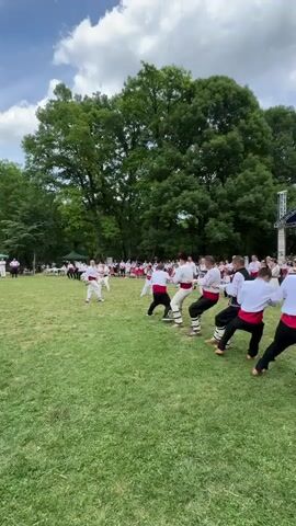 A group of people in traditional clothing are playing tug-of-war on a grassy field. The players are divided into two teams, each wearing white shirts with red sashes and black pants. The field is surrounded by trees and a blue sky with white clouds. There is a stage in the background with some people on it. The game is clearly in progress with both teams pulling on a rope, indicating a moment of intense effort.