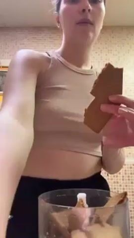 A young woman stands in a kitchen, breaking pieces of gingerbread into a food processor. She has her hair up, is wearing a tan tank top, and black sweatpants. The kitchen has tan tile in the background and what looks like an air conditioner.