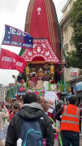 A religious procession is taking place on a street. In the center of the frame is a large, ornate chariot decorated with red and green fabric, topped with a white cross symbol. Several people are visible on the chariot, some appearing to be tending to statues or deities. In front of the chariot, a crowd of people is gathered, watching the procession. Some individuals in the crowd are holding flags, including one that says "JESUS IS OUR ONLY HOPE" and another that says "JESUS IS KING." The environment appears to be an urban street with buildings lining one side of the road and trees in the background. The sky is overcast. The video is shot from the perspective of someone in the crowd, looking towards the chariot.