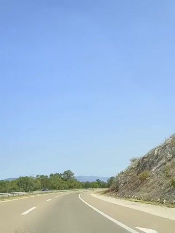 The video shows a view from a moving vehicle on a clear, sunny day. The camera is pointed forward and slightly to the right, capturing a highway with two lanes of traffic going in the same direction. On the left side of the road, there is a median strip with some vegetation, and beyond that, more trees and distant hills. On the right side of the road, there is a rocky embankment covered in dry brush and a low fence. The sky is a bright, clear blue with a few faint smudges that could be lens flare or dirt on the camera lens. The road appears to be paved with asphalt and has white lane markings. There are no people visible in the video, and no other vehicles are close enough to be distinctly identified. The overall impression is of a scenic drive through a somewhat arid landscape.