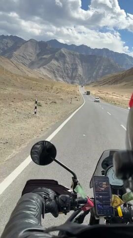 A first-person perspective video shows a motorcycle rider on a paved road surrounded by barren, mountainous terrain under a cloudy sky. The rider, wearing black leather gloves and a black leather jacket, grips the motorcycle's handlebars. A smartphone is mounted on the handlebars, displaying a navigation app. Prayer flags are attached to the motorcycle. Several vehicles, including a truck and cars, are visible on the road ahead, heading in the same direction. The landscape is characterized by rugged, eroded mountains and dry, sandy ground.