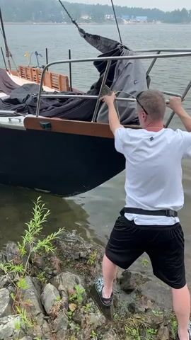 A man in a white t-shirt and black shorts stands on a rocky shore, holding onto the side of a docked sailboat. The sailboat has a black hull with white lettering and a drawing of a figure. Several people, including children wearing life vests, are sitting on the deck of the sailboat. The water is calm and the sky is overcast. In the background, there are trees and buildings on a distant shore.