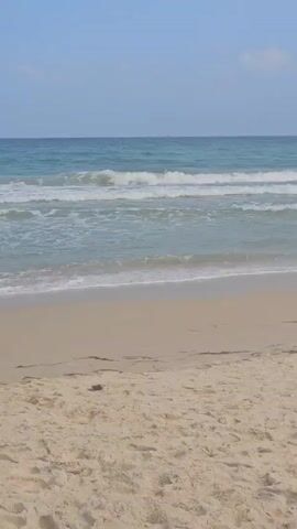 The video shows a sandy beach on a partly cloudy day. The sand is light-colored, and there are some bits of debris scattered on it. The ocean stretches out to the horizon with small waves gently lapping at the shore. In the distance, a few buoys or markers are visible. The sky is a mix of blue and scattered clouds. There are no people visible in the frame. The camera is static, providing a wide shot of the beach and ocean.