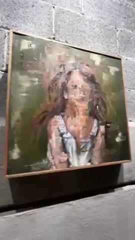 The video shows a series of paintings displayed on a gray cinder block wall. The paintings are framed in a light-colored wood and appear to be portraits in a blurred, abstract style. The first painting is of a girl in a white dress with a green and brown background. The second painting is of a person in a dark jacket with a light blue background. The third painting is of a person sitting on a stool. A piece of paper with green text and a graph is also visible on the wall. The camera pans up and across the wall, showing the artwork at different angles. The environment appears to be an art gallery or studio with a rough, industrial feel. There are no people visible in the video.