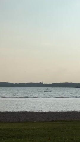 A person is paddleboarding on a lake. They are in the middle of the lake, a good distance from shore. The lake is wide and calm, with a faint ripple on the surface. The shore is lined with trees. The sky is a pale blue with a few wispy clouds. In the foreground, there is a grassy area with some pebbles. The overall impression is one of peace and tranquility.