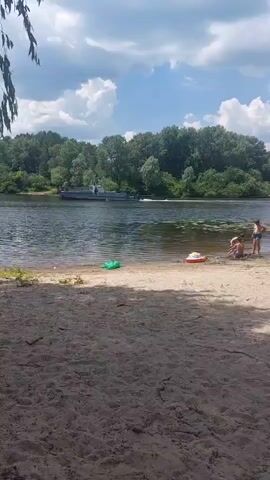 The video shows a sandy beach with a river in the background. The river is lined with trees and there is a boat on the water. There are three people on the beach, two adults and one child, wearing swimwear. One of the adults is sitting on the sand, while the other adult and child are standing near the water's edge. A red and white inflatable ring is floating in the water. There is also a green inflatable object on the sand. The sky is partly cloudy.