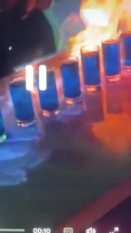 A row of shot glasses filled with a blue liquid is set ablaze on a reflective surface, possibly a bar or counter. Flames engulf the top of the shot glasses, casting a warm orange and yellow glow on the scene. The background is dark and blurred, with hints of green and blue lighting. The camera is positioned at a slight downward angle, focusing on the burning drinks.