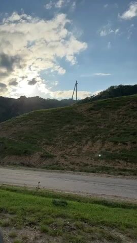The video shows a landscape with a road, a grassy hill, and mountains in the background. The sky is blue with some clouds. There are a few telephone poles visible on the hill. The camera pans across the scene, showing different angles of the landscape. The road appears to be paved and is bordered by grass. The hill is covered in grass and some bare patches of dirt. The mountains are green and appear to be covered in trees. In the distance, there is a small village visible in the valley between the hills. The video captures a peaceful and scenic view of a rural area.