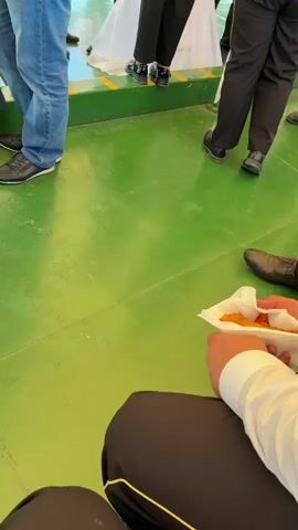 The video shows a view from a boat on a sunny day. The camera pans up from a person holding a snack to show the interior of the boat, filled with people, and then shifts to the exterior view of a wide river bordered by trees. The sky is clear blue. The boat has a yellow canopy.