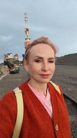 A woman with short, blonde hair and makeup is standing in the foreground, facing the camera. She is wearing an orange cardigan over a pink t-shirt and has a backpack on. In the background, there is a ship docked at a pier, a large industrial crane, and a pile of gravel. The sky is cloudy. The woman is looking at the camera and smiling slightly. The scene appears to be a port or industrial area.