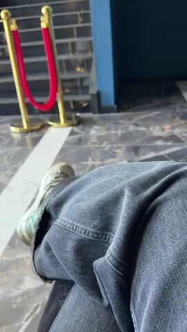 The video shows a person sitting, wearing dark denim pants and light green sneakers, in what appears to be a lobby or waiting area. There's a golden stanchion with a red rope visible in the background. The camera quickly pans to the right, showing a hallway with blue walls and mirrors, and then focuses on a sign that reads "MAESTRO HALL" in white lettering on a blue wall. There is also a stylized 'M' logo next to the hall's name and a wheelchair accessible icon.