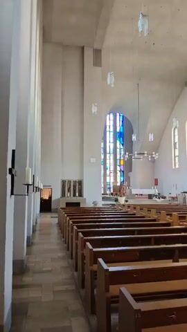 This is a video showing the interior of a modern church in Memmingen, Germany. The scene is captured from a low angle, looking towards the altar. The church has a large, open nave with rows of wooden pews on either side. The walls are smooth and off-white, with tall, arched windows that let in natural light. Above, the ceiling is high and curved, with several modern pendant lights hanging down.

A large, colorful stained-glass window is visible at the far end of the church, behind the altar. The altar itself is simple, with a white cloth draped over it. There are some religious symbols and artwork visible on the walls and near the altar. The overall impression is one of spaciousness and contemporary design. There are no people visible in the video, suggesting it was filmed during a quiet time, perhaps before or after a service, or when the church is closed to the public. The lighting suggests it is daytime. The atmosphere appears peaceful and serene, befitting a place of worship. No text is visible in the media file.