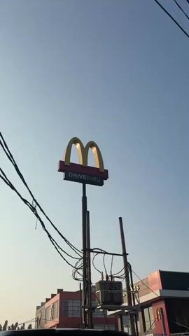 This is a vertical video of a bright, clear sky with a few clouds. In the foreground, there is a McDonald's sign with "DRIVE THRU" written below it. The sign is attached to a tall pole with electrical wires. To the left of the McDonald's sign, there is a red building with multiple floors and large windows. To the right of the McDonald's sign, there is another building with the McDonald's logo and text that appears to say "McDonald's Indonesia". Further to the right, there is a Domino's Pizza sign. The video pans slightly to the right, revealing more of the buildings and surrounding area, including trees and a street light. The overall impression is of a commercial area on a sunny day.