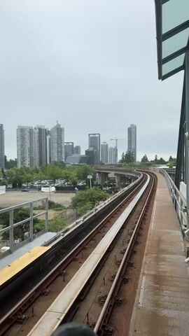 The video pans across a railway track and its surrounding environment. In the foreground, the tracks are visible, with one set of tracks curving to the right. To the right of the tracks is a raised platform with a weathered, metallic surface. A portion of a glass and metal canopy structure is visible at the top right of the frame.

In the mid-ground, the railway line continues its curve, passing under a concrete overpass. Trees with lush green foliage line the area beside the tracks and beneath the overpass. A few cars are parked in a lot to the left of the overpass. A train is seen approaching and traveling along the overpass.

In the background, a cityscape unfolds with numerous tall buildings of varying heights and architectural styles. A construction crane is visible among the buildings. The sky is overcast, suggesting a cloudy or possibly rainy day. There are a few indistinct figures walking on a path or sidewalk below the overpass. The overall scene depicts a transit infrastructure within an urban setting on a cloudy day.
