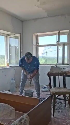 A man is mixing cement in a bucket using a power drill in a room under renovation. The room has two windows looking out on a landscape with trees. The room's ceiling has an exposed lightbulb. There's a wooden dresser, a plastic water bottle, and bags of cement on the floor. There's a table with tools and a broken chair in the room. The walls are freshly plastered.