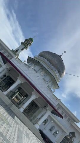 This video features an eye-level, slightly upward-angled shot of a white mosque with a prominent silver-colored dome and a tall, slender minaret. The mosque, identified as Mesjid Reubee in Indonesia, showcases traditional Islamic architecture with intricate patterns on its facade, white pillars, and decorative railings. The dome has a crescent moon and star finial at its apex, as does the green-capped minaret.

The foreground shows a paved courtyard with grey and white tiles, some arranged in a circular decorative pattern. The sky above is a bright blue with scattered wispy white clouds, indicating clear weather during the daytime, likely morning or early afternoon due to the lighting. No people or activities are visible, emphasizing the mosque's peaceful and grand presence. The video is a static shot, capturing the architectural beauty of the religious building against a serene sky.
