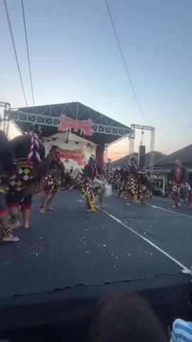 A vertical, low-resolution video displays an outdoor cultural performance. The scene is dominated by a raised stage with a black floor, set against a backdrop of a white and red banner and stage lighting. Several performers, adorned in traditional Indonesian attire, including patterned sashes, head coverings, and what appear to be horse or animal headdresses, are actively engaged in a dance. The performers are moving across the stage, some appearing to be in mid-stride or leap. Petals are scattered on the stage floor, possibly from the performance.

In the foreground, the edge of a stage is visible, with a dark surface. Behind this, a large crowd of spectators is gathered, also in vertical orientation, watching the performance. The audience comprises people of various ages, dressed in casual clothing. Some are looking towards the stage, while others are conversing or holding up phones, likely to record the event. The environment suggests a warm climate, with a clear sky transitioning to dusk. Buildings with pitched roofs are visible in the background, as are power lines and a small tower with a speaker or light fixture. The overall atmosphere appears to be festive and community-oriented.