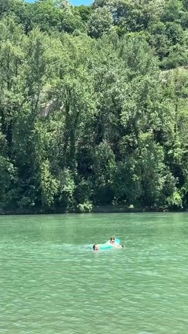 The video shows two people in a large body of green water. One person is lying on a light green inflatable float, and the other person is swimming beside them. The water is calm, and there are trees on the bank in the background, with a clear sky above. The scene appears to be a natural outdoor environment on a sunny day.