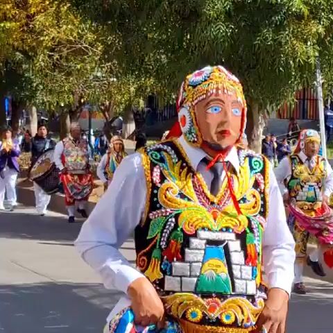 A parade is taking place on a paved street lined with trees. The participants are dressed in ornate, colorful traditional costumes. The costumes include headdresses, masks with painted faces, vests adorned with intricate embroidery and geometric patterns, white shirts, ties, and trousers. Some participants are wearing red scarves. One participant in the foreground has a vest featuring a depiction of a stepped pyramid with a sun symbol. Several participants are holding or wearing items that appear to be part of their performance, such as colorful sashes and skirts. In the background, other parade participants, musicians with drums, and onlookers can be seen. The environment suggests a daytime outdoor event, possibly in a town or city, given the buildings visible on a hillside in the distance and streetlights. The overall atmosphere seems festive.
