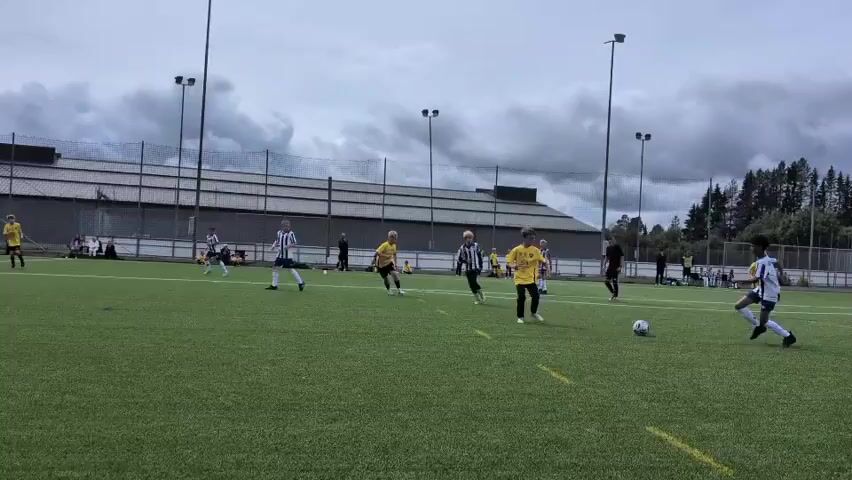 The video shows a soccer game being played on an artificial turf field. There are two teams, one in yellow jerseys and one in white and black striped jerseys. The players are running and kicking the ball. There are spectators in the background sitting on benches. There are tall light poles around the field. The sky is cloudy. The field has yellow lines marking the boundaries. There is a blue flag with a logo on it in the foreground.