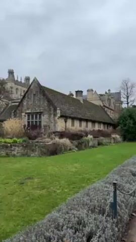 The video shows a view of a stone building and green lawn under a cloudy sky.  The building in the foreground is long and low with a steeply pitched roof. Behind it is a taller stone structure with a tower. A manicured lawn stretches out in front, bordered by a low hedge.  The scene is still and peaceful, suggesting a location in a historical or academic setting.