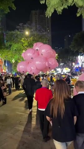 The video depicts a crowded street at night, with people walking among the illuminated buildings and trees. In the foreground, a cluster of pink, pig-shaped balloons floats above the crowd. Many pedestrians wear casual clothing, and the atmosphere seems lively and festive. There's a mix of individuals and groups moving in various directions. The street is well-lit, casting long shadows, and the overall scene captures an urban nightlife environment.