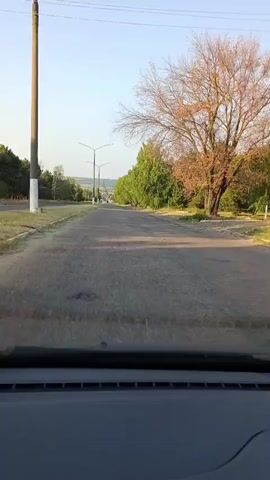 The video shows a view from inside a vehicle driving down a paved road. The road is lined with trees and streetlights. The sky is clear and the sun is shining. The trees on the right side of the road are tall and green, with some appearing to be deciduous and losing their leaves. The trees on the left side are also green, and appear to be evergreen. The road itself appears to be in moderate condition, with some cracks and discoloration visible. There is a grassy area on the left side of the road, which is also lined with trees. The camera is focused on the road ahead, and the vehicle is moving forward. The overall impression is of a rural or suburban setting, possibly in the early morning or late afternoon, given the lighting.
