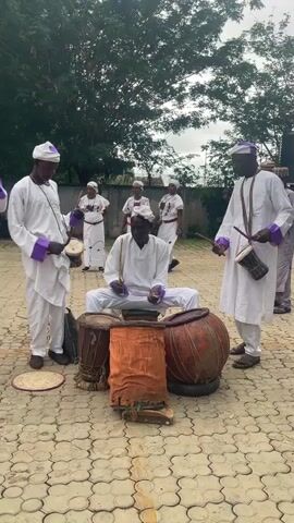 A video clip features a group of people dressed in white clothing with purple accents, participating in a drumming performance outdoors. The central figure, a man sitting cross-legged, is actively playing a large drum placed in front of him. To his left and right stand two other men, also in white attire, each holding and playing a smaller drum. Behind them, a group of women, similarly dressed, observe the performance. The ground is paved with interlocking bricks, and a lush green tree provides a natural backdrop. The overall atmosphere suggests a cultural or ceremonial event.