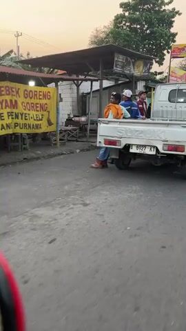 In Kedungcangkring, Indonesia, a white Suzuki pickup truck is parked on a paved road. Three people are seated in the truck bed. The person closest to the camera is wearing an orange shirt and jeans. The person behind them is wearing a blue jacket and a white cap. The third person is wearing a red and white striped shirt and a dark cap. A yellow banner is hanging from a canopy on the left side of the road. The banner has text in Indonesian. The truck has a license plate that reads "A 8927 KT". The scene appears to be outdoors during the day, possibly late afternoon given the lighting. The sky is a pale orange, suggesting the sun is low on the horizon. There are some buildings and trees visible in the background. The overall impression is of a casual, everyday scene in a semi-rural or roadside setting. The people in the truck appear to be relaxed.