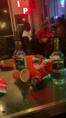 The video pans across a table covered in discarded red plastic cups, empty aluminum cans, and two bottles of Jameson Black Barrel whiskey. In the background, four people are seated at a table. One man in a white shirt is on his phone. A man in a red shirt is talking to a woman in a dark top. The environment appears to be a dimly lit indoor venue with neon signage that reads "VIP." The lighting is predominantly red and blue.