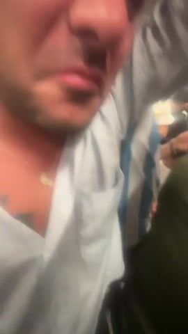 A close-up, shaky video shows a man's face as he makes a disgusted or upset expression, puckering his lips and furrowing his brow. He is wearing a light blue collared shirt with thin white pinstripes and a necklace with a small pendant. A tattoo is partially visible on his chest. The background is blurry but suggests an indoor setting with other people present, one of whom appears to be wearing a white and blue striped garment. The lighting is warm and somewhat dim.