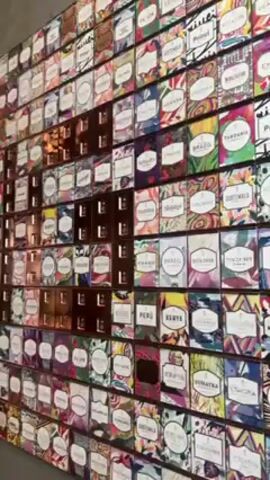 The video shows a large display on a wall made up of numerous small, colorful boxes. Each box has a label on it, and the labels appear to indicate different countries of origin, such as Brazil, Colombia, and Kenya. Some of the boxes have copper-colored, recessed handles. The wall appears to be made of concrete. In the background, there are a few people sitting at tables. The camera slowly pans away from the wall to give a wider view of the scene.