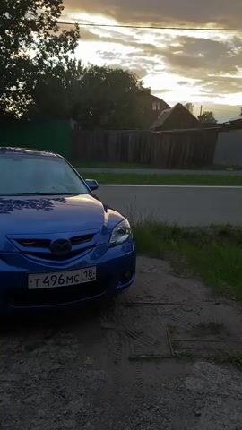 A blue Mazda 3 is parked on a gravel and dirt area next to a road. The car is angled towards the viewer, with its front end and hood visible. The car has a Russian license plate that reads "T 496 MC 18 RUS". In the background, there is a road, a wooden fence, some trees, and houses. The sky is cloudy with the sun setting, casting a warm glow on the scene. The overall atmosphere is peaceful and serene. The time of day is sunset, and the weather appears to be clear with some clouds. The location is Votkinsk, Russia, as indicated by the license plate.