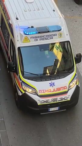 A high-angle shot shows the rear of a white ambulance with blue flashing lights on the roof. The ambulance has "MISERICORDIA DI PESCIA" written on its side, along with a Star of Life symbol and the text "AMBULANZA". The license plate reads "CZ 107DJ". The ambulance appears to be stopped on a street with a cobblestone sidewalk. A lamppost and a potted plant are visible on the sidewalk. A small, awning-covered structure is partially visible on the left side of the frame. Reflections of buildings are visible on the ambulance's windshield.