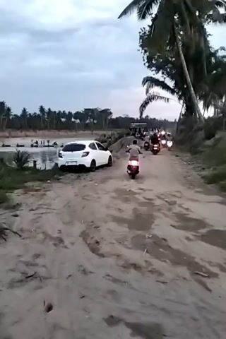 The video shows a dirt road beside a body of water, likely a river. A white hatchback car is parked on the left side of the road, close to the water's edge. On the right side, a line of people on motorcycles are moving towards the camera on the dirt road. Several coconut palm trees are visible in the background, and the sky is overcast. People can be seen near the water in the background as well.