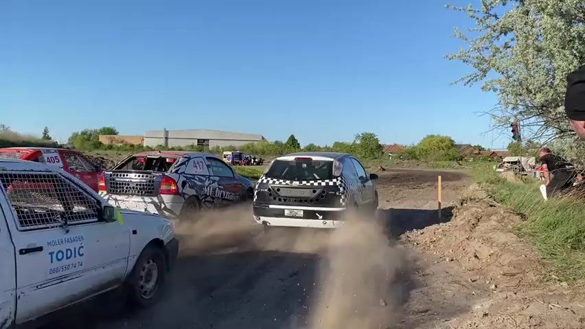 The video shows four rally cars racing along a dusty, unpaved track. The cars are of different colors and models, and each kicks up a large cloud of dust. A man wearing a black beanie watches from the side of the track with some other spectators. The scene is outdoors on a sunny day, with a clear blue sky and trees in the background. The presence of a building in the distance suggests a rural or semi-rural environment.