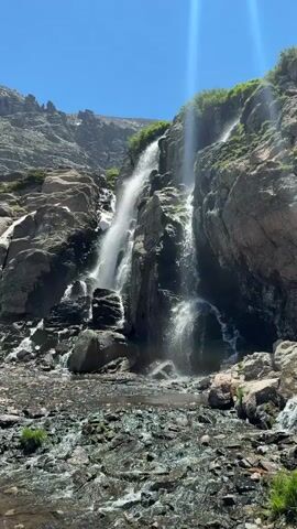 This is a vertical video of a waterfall cascading down a rocky mountainside. The water is white and frothy as it falls, creating a mist at the bottom. The rocks are dark gray and rough, with some patches of green vegetation growing on them. The sky above is clear and blue. The video is filmed from a low angle, looking up at the waterfall. The sun is shining brightly, and there are lens flares visible in the upper right portion of the frame. The bottom of the frame shows a streambed filled with rocks and some water.
