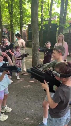 An outdoor scene features approximately a dozen children and several adults gathered in a wooded area for what appears to be a laser tag activity. The ground is dirt, and numerous tall trees are visible. Most individuals, primarily children, are equipped with laser tag headbands containing sensors and are holding various models of laser tag guns. In the foreground, a girl in a light blue t-shirt and colorful shorts smiles while holding a gray laser tag gun, looking towards another girl in a dark t-shirt who is holding a larger black laser tag gun. A boy in a green t-shirt stands between them, also wearing a headband and holding a gun. Several other children and adults are scattered in the midground and background, similarly equipped with laser tag gear. An adult woman in a white dress stands near a blue table in the left background, while another adult woman in light attire stands in the right background. Participants are engaged in conversation and interaction.