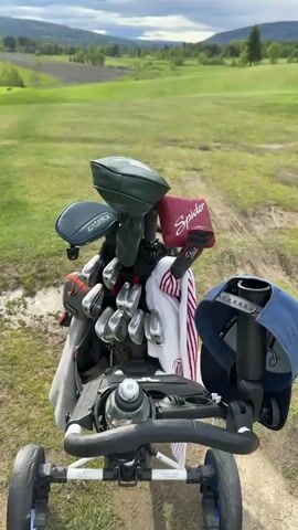 The video opens with a close-up shot of a golf bag and push cart. The golf bag is packed with various clubs, each with its head cover. There's also a towel and a water bottle attached to the cart. The background reveals a picturesque golf course with rolling green hills and scattered trees under a cloudy sky.

The camera then pans across the green, showing the well-manicured grass. The view shifts to a tee box where two men are playing golf. One man in dark trousers and a blue cap is observing the other. The second man is wearing a blue shirt and shorts, and he is positioned to swing at a golf ball. A golf bag is placed on a stand next to a wooden bench in the foreground.

The final scene provides a wider view of the golf course, showcasing its hilly landscape and scattered trees. The sky is a mix of clouds and patches of sunlight. There are two people walking on the green in the distance.