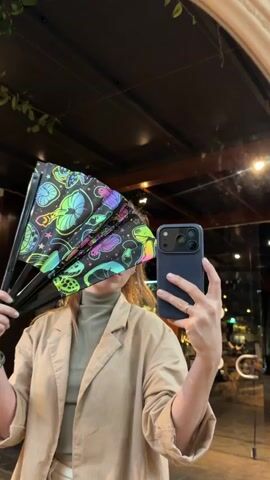 A woman is filming herself in a mirror or reflective surface, holding a large, open folding fan. She wears a light-colored top, possibly beige or cream. Her left hand holds a dark-cased smartphone (appears to be an iPhone) with its camera facing her, suggesting she is recording a selfie video. Her right hand holds the fan.

The fan is black with a vibrant, multicolored mushroom pattern, featuring various shapes and sizes of mushrooms in neon greens, pinks, purples, blues, and oranges. The woman looks directly at her phone's camera, with a neutral to slightly smiling expression. She has light hair pulled back. A ring is visible on her right hand.

The background, seen through the reflection, suggests an indoor or covered outdoor setting with dim lighting. Elements like hanging plants, exposed ceiling structures, and small lights are visible. The overall ambiance appears to be a relaxed evening setting, possibly in a restaurant or bar in Nir Eliyahu, Israel.