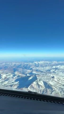 This media file captures an aerial view of snow-capped mountains under a clear blue sky. The perspective is from inside an aircraft, looking out over a vast mountain range. The foreground shows a portion of the aircraft's dashboard or cockpit, with a row of small black indentations along its edge.

The mountains themselves are rugged and appear to be at a high altitude, with patches of snow contrasting with exposed rocky surfaces. The landscape stretches into the distance, with layers of smaller mountain ranges fading into a hazy horizon. The bright sunlight suggests it is daytime, likely late morning or early afternoon, given the clear visibility and the illumination of the terrain.

The overall impression is one of majestic natural beauty and the awe-inspiring scale of mountain landscapes from above. The clear sky indicates fair weather. While the exact location is not explicitly stated in the visual, the presence of snow-capped mountains suggests a region with significant elevation and cold temperatures, common in alpine or high-altitude environments. There is no visible text in the media file.