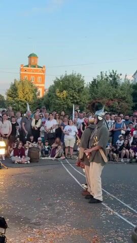 A group of people in what appears to be a historical reenactment, dressed in what looks like Roman legionary armor, are standing in formation in the middle of a street. They are facing a crowd of spectators who are seated and standing on the side of the street. The crowd is diverse in age and gender, and many are holding up their phones to record the event. In the background, there is a tall, orange building with a green dome, surrounded by trees. The sky is clear and the sun is setting, casting a warm glow on the scene. There are also some stage lights set up on the left side of the frame, indicating a performance is taking place. Confetti is scattered on the ground in front of the performers and the crowd.
