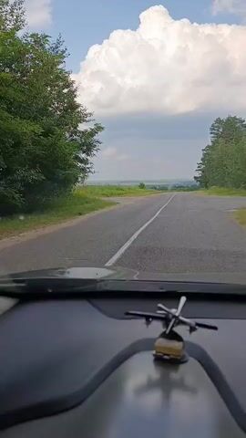 The video captures a point-of-view shot from inside a car as it travels along a rural road. The road is paved with a white line marking the center. On either side of the road are grassy verges and trees, with a more dense forest on the right. In the distance, the landscape opens up to a wide, hazy view of fields and a distant body of water or plain under a bright, cloudy sky. A small, metallic helicopter figurine is visible on the dashboard in the foreground, slightly out of focus.

The activity is driving on a road, suggesting travel or a journey. The time of day appears to be daytime, likely late morning or afternoon, given the bright sunlight and the formation of the clouds. The weather seems fair with dramatic, fluffy white clouds against a blue sky, indicating a possibility of changing weather or a generally picturesque day. The setting is rural, with a focus on the natural landscape and a sparsely populated road. The location context provided, Козенскі сельскі Савет, Belarus, situates this scene within the Belarusian countryside.

There is no visible text in the video. The overall mood conveyed is one of peaceful travel and observation of the scenery. The drone-like perspective of the road stretching into the distance and the open sky contribute to a sense of freedom and exploration.