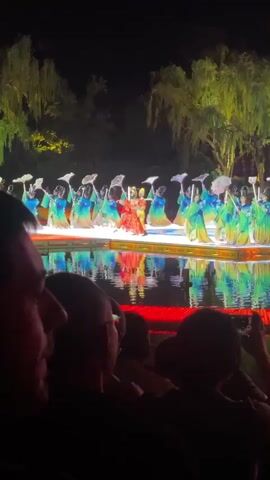 A video captures a nighttime performance on a stage set over water, observed by a seated audience. The stage is brightly lit, reflecting colors onto the water's surface. The backdrop features what appear to be willow trees, enhancing the theatrical atmosphere. Performers, dressed in vibrant costumes ranging from yellow to red, blue, and green, are engaged in a choreographed dance. They utilize props such as parasols and long-handled instruments, moving in unison. The audience, primarily visible from the back, consists of people of various ages and genders, some of whom are recording the performance with their phones. The focus remains on the stage performance, with the audience serving as an observing presence.