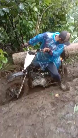 The video shows a man on a motorcycle stuck in thick mud in a forested area. He is wearing a blue poncho and a baseball cap. Another man, also wearing a poncho, helps him by pushing a large log across the motorcycle's back tire, apparently to assist with traction. The environment is lush with dense green vegetation.
