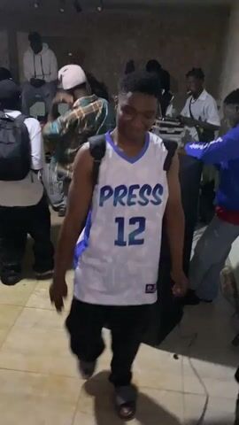 A young Black male in a white basketball jersey with "PRESS 12" in blue lettering is dancing and holding a phone. He is wearing black pants and gray sandals. He is in an indoor setting with other people in the background. There is a DJ setup with speakers and a laptop to the right of the frame.