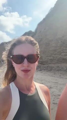 A young woman with blonde hair and sunglasses is standing on a sandy beach. She is wearing a green and white swimsuit. The ocean and a cliff face are visible in the background. The sky is blue with some clouds.